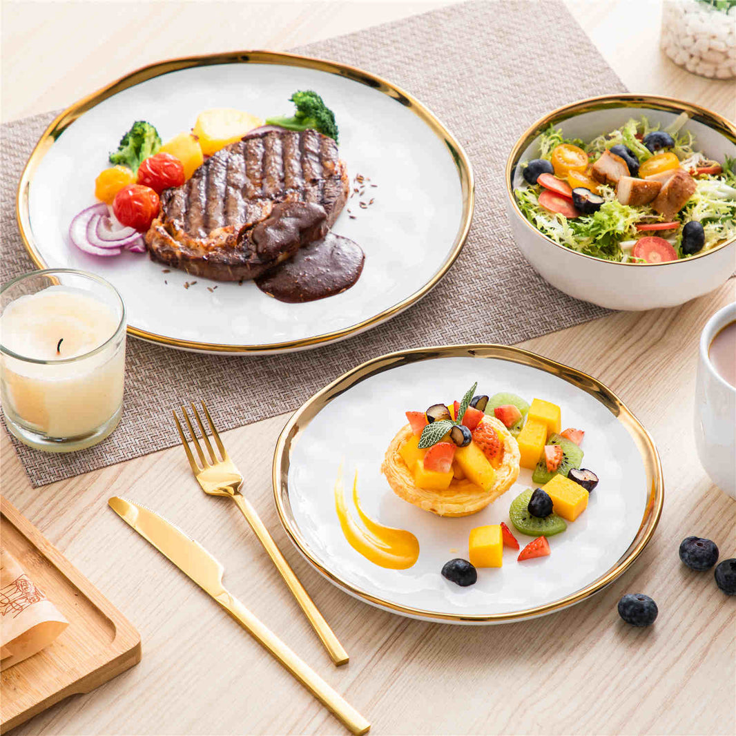Lovecasa 16-piece white porcelain dinnerware set with gold trim, styled for solo dining with steak, tart, salad bowl, and candle on a cream table setting.
