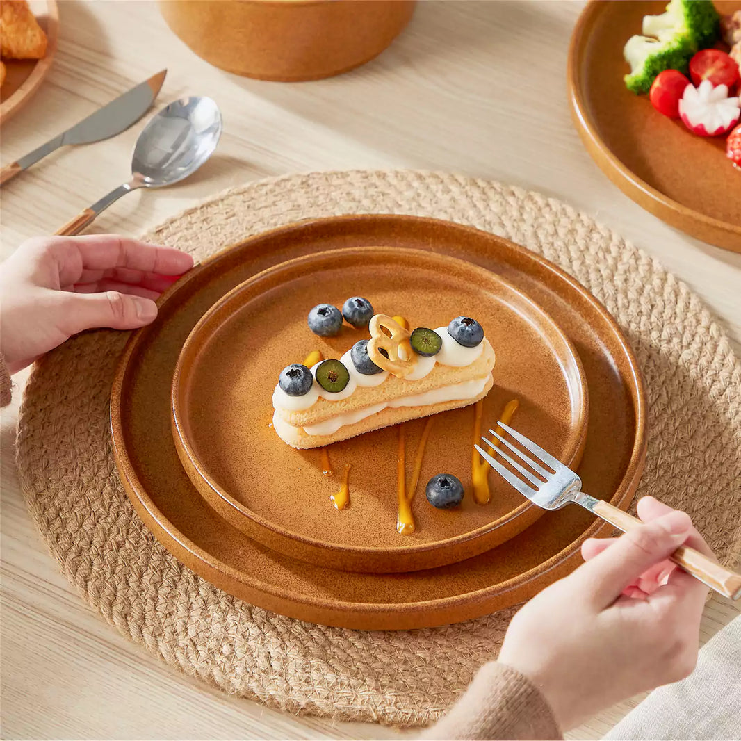 Webb Straight-Edge caramel reactive glaze stoneware dinner and salad plates stacked on woven placemat with blueberry cake solo dining setup on off-white wood table.